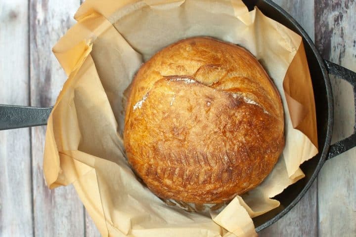 A round loaf of golden brown bread sits on parchment paper inside a black cast iron pot.