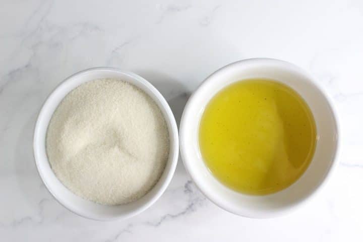 Two white bowls on a marble surface, one filled with granulated sugar and the other with egg whites.