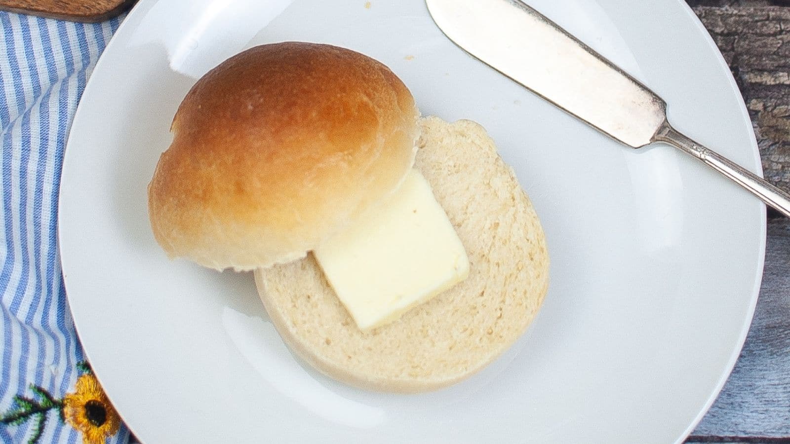 A bread roll sliced in half with a pat of butter inside, placed on a white plate next to a butter knife.