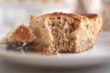Close-up of a partially eaten slice of apple sour cream coffee cake with visible apple pieces, resting on a white plate with a fork holding a bite-sized piece.