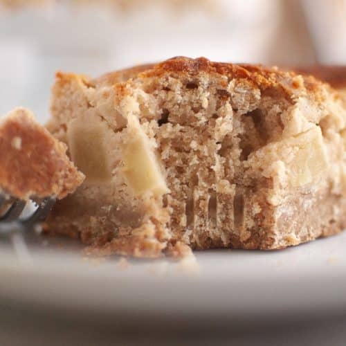 Close-up of a partially eaten slice of apple sour cream coffee cake with visible apple pieces, resting on a white plate with a fork holding a bite-sized piece.