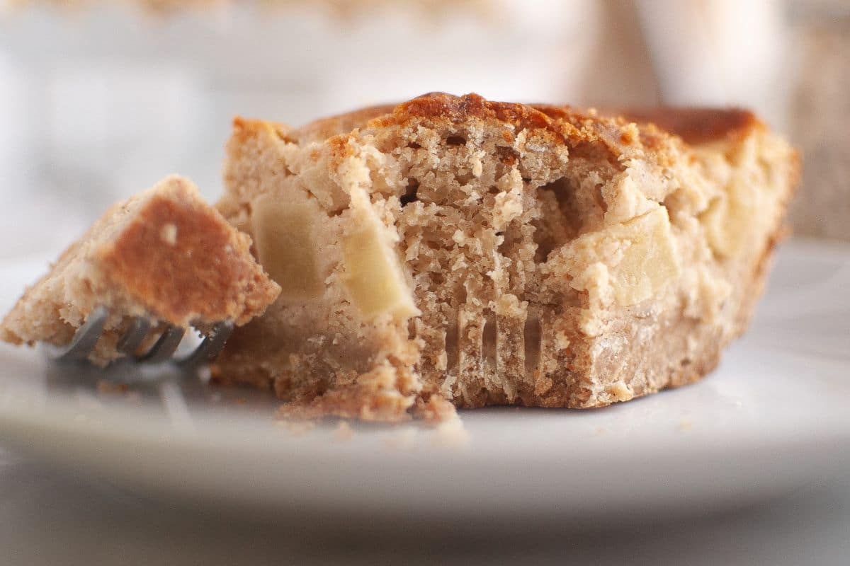 Close-up of a partially eaten slice of apple sour cream coffee cake with visible apple pieces, resting on a white plate with a fork holding a bite-sized piece.