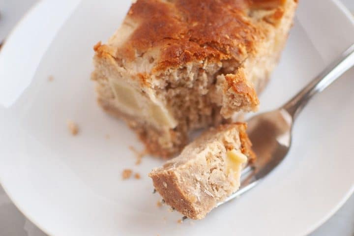 A slice of crumbly apple sour cream coffee cake with visible apple pieces on a white plate, with a fork holding a bite-sized piece.
