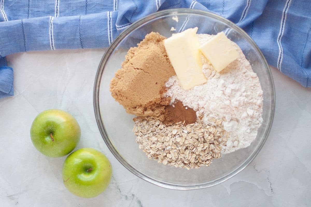 A glass bowl contains brown sugar, oats, flour, cinnamon, and two butter sticks on a marble surface next to two green apples and a blue cloth.