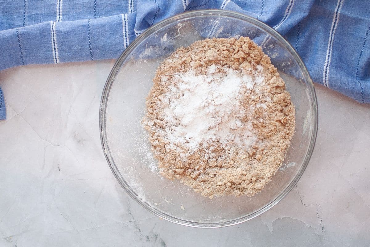 A glass bowl with flour and brown sugar mixture, topped with baking powder, sits on a marble surface next to a blue striped kitchen towel.