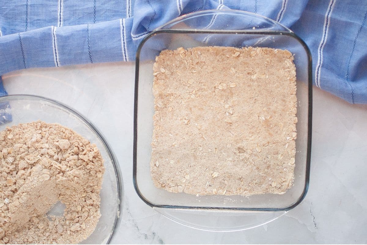 A glass baking dish with an even layer of oat and flour mixture sits on a marble surface beside a bowl of the same mixture and a blue striped cloth.