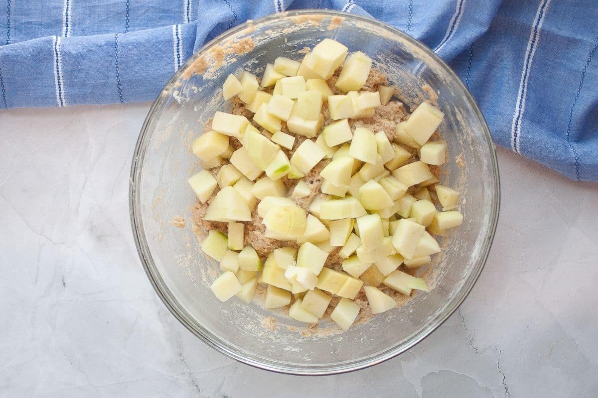 A glass bowl filled with chopped apples mixed into a batter, placed on a light marble surface next to a blue striped kitchen towel.
