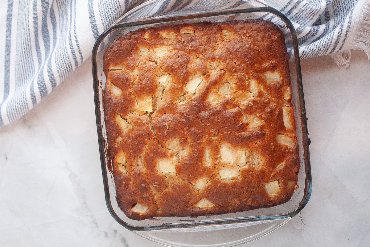 A square glass baking dish filled with a golden-brown cake containing visible apple chunks sits on a white surface next to a striped cloth.