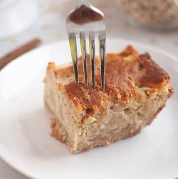 A fork is pressed into a slice of apple cake on a white plate, with a cinnamon stick and a striped napkin nearby.