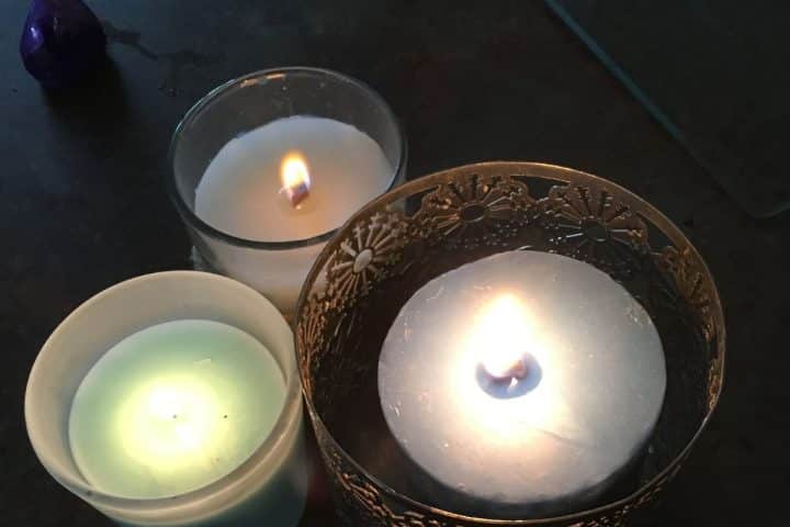 Three lit candles in glass holders are arranged close together on a dark surface. One candle is white, one is blue, and one is pale green.