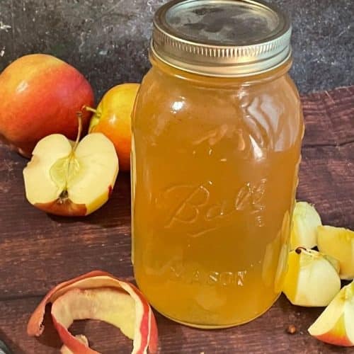 A glass mason jar filled with amber liquid sits on a wooden surface, surrounded by whole and sliced apples, apple peels, and a peeler.