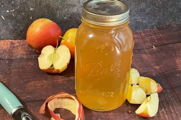 A glass mason jar filled with amber liquid sits on a wooden surface, surrounded by whole and sliced apples, apple peels, and a peeler.