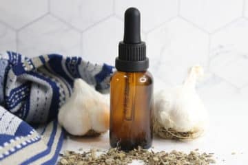 A brown glass dropper bottle of mullein garlic oil for ear aches sits between two heads of garlic, with dried herbs and a blue-striped cloth nearby on a white surface.