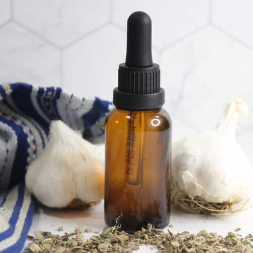 A brown glass dropper bottle of mullein garlic oil for ear aches sits between two heads of garlic, with dried herbs and a blue-striped cloth nearby on a white surface.