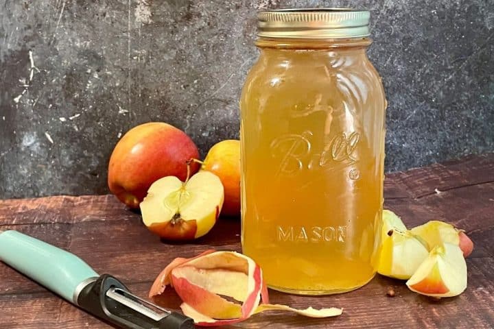 A jar of light amber liquid sits next to sliced apples, apple peels, a paring knife, and whole apples on a wooden surface.