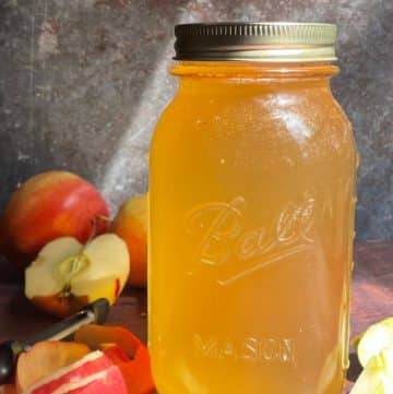 A mason jar filled with golden homemade apple cider vinegar sits on a wooden surface, surrounded by whole and sliced apples and a corer.