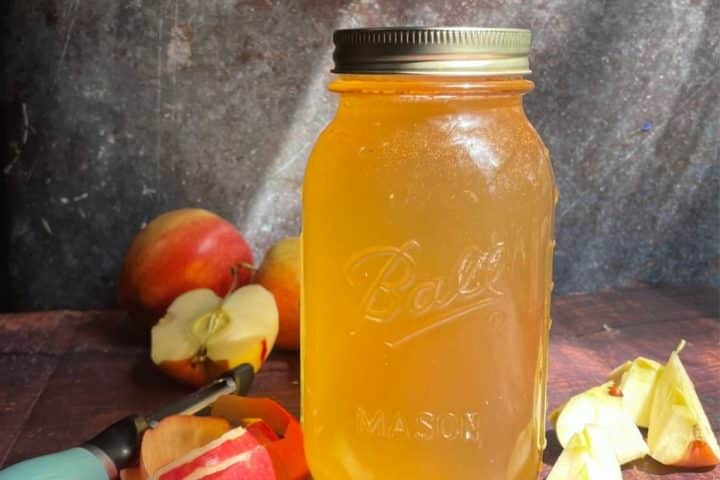 A mason jar filled with golden homemade apple cider vinegar sits on a wooden surface, surrounded by whole and sliced apples and a corer.