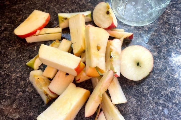 Sliced and cored apple pieces are scattered on a dark countertop next to an empty glass jar.