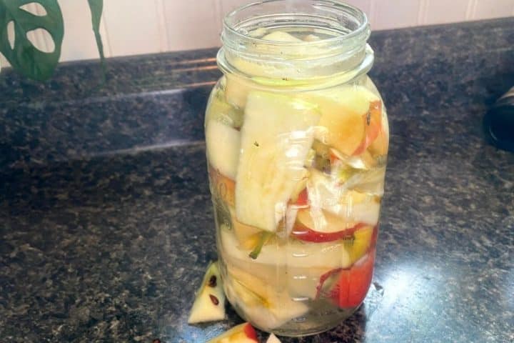 A glass jar filled with sliced apples and other fruit pieces soaking in liquid, placed on a dark kitchen countertop.