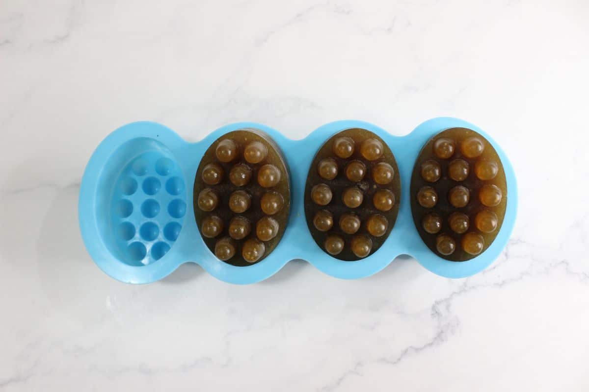 A blue silicone mold holds three brown oval-shaped soaps with raised bumps, placed on a white marble surface.