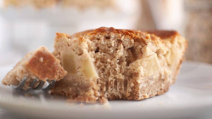 A slice of apple sour cream coffee cake with a bite taken out, sitting on a white plate next to a fork with a piece of cake on it.