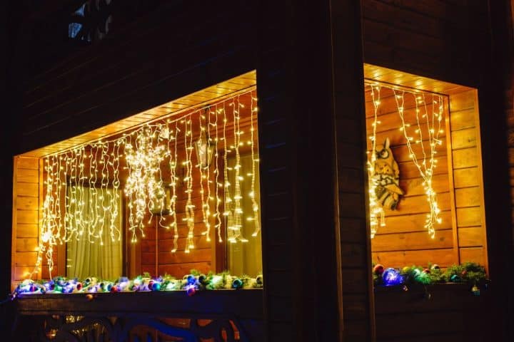 A wooden house with a large window decorated with hanging string lights and a garland of colorful lights along the windowsill.