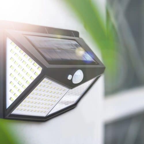 A wall-mounted outdoor solar-powered LED light with a motion sensor, installed beside a window, illuminated by sunlight.