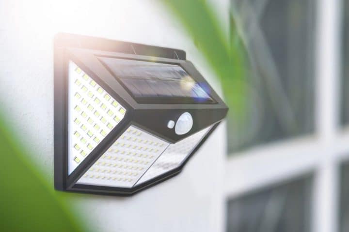 A wall-mounted outdoor solar-powered LED light with a motion sensor, installed beside a window, illuminated by sunlight.