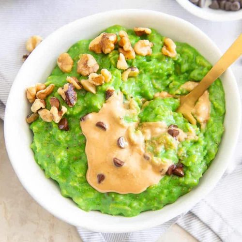 A bowl of bright green oatmeal makes for a filling, cold-weather breakfast, topped with peanut butter, chopped walnuts, and chocolate chips, with a gold spoon beside it.