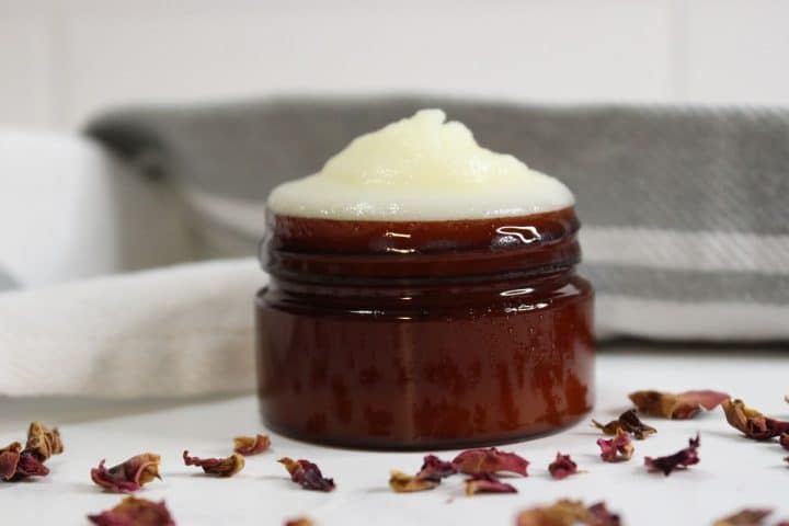 A small brown jar filled with a creamy white substance, surrounded by dried flower petals, sits on a white surface with a gray cloth in the background.