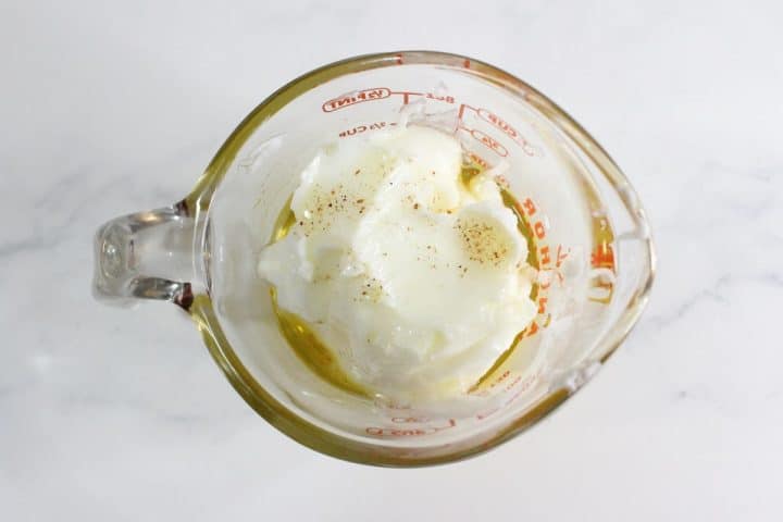 A glass measuring cup containing mayonnaise, oil, and black pepper on a white surface.