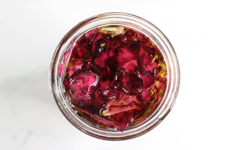 A glass jar filled with red or purple flower petals submerged in a liquid, viewed from above against a white background.