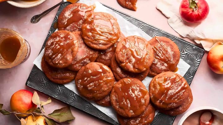 A tray of caramel-covered apple cookies on a patterned plate, surrounded by apples and a glass of caramel sauce.
