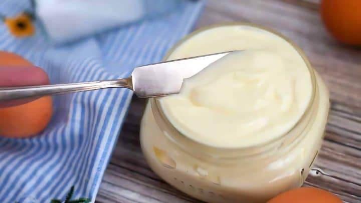 A knife spreads creamy mayonnaise from a jar on a wooden surface, with oranges and a striped cloth nearby.