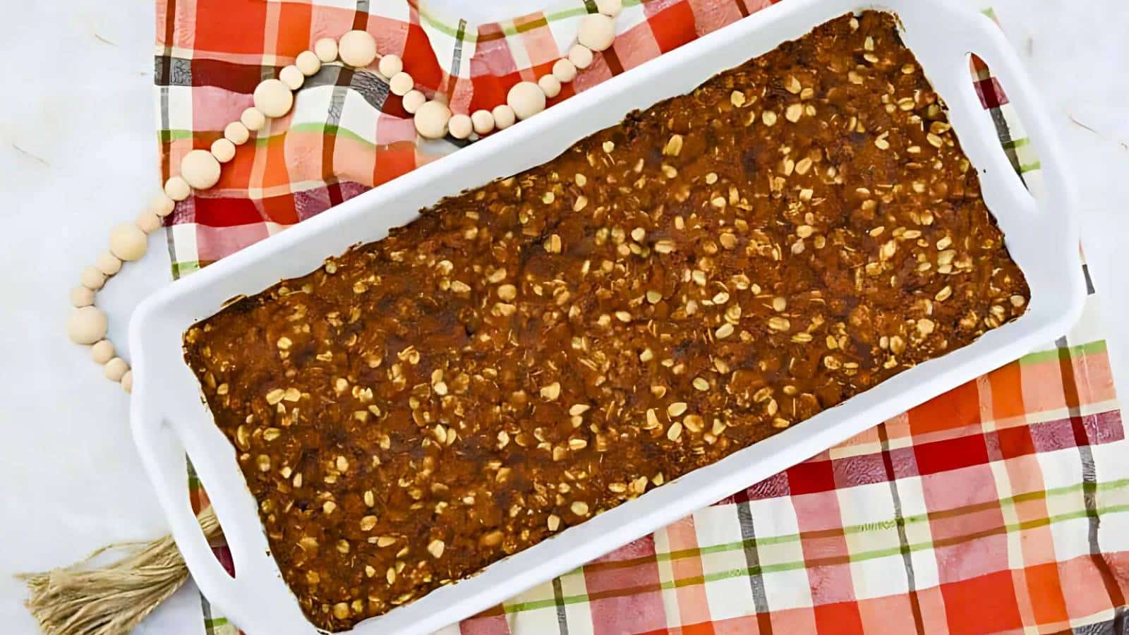 A rectangular baked dish with oats and nuts on a white tray, placed on a colorful plaid cloth beside a wooden bead garland.