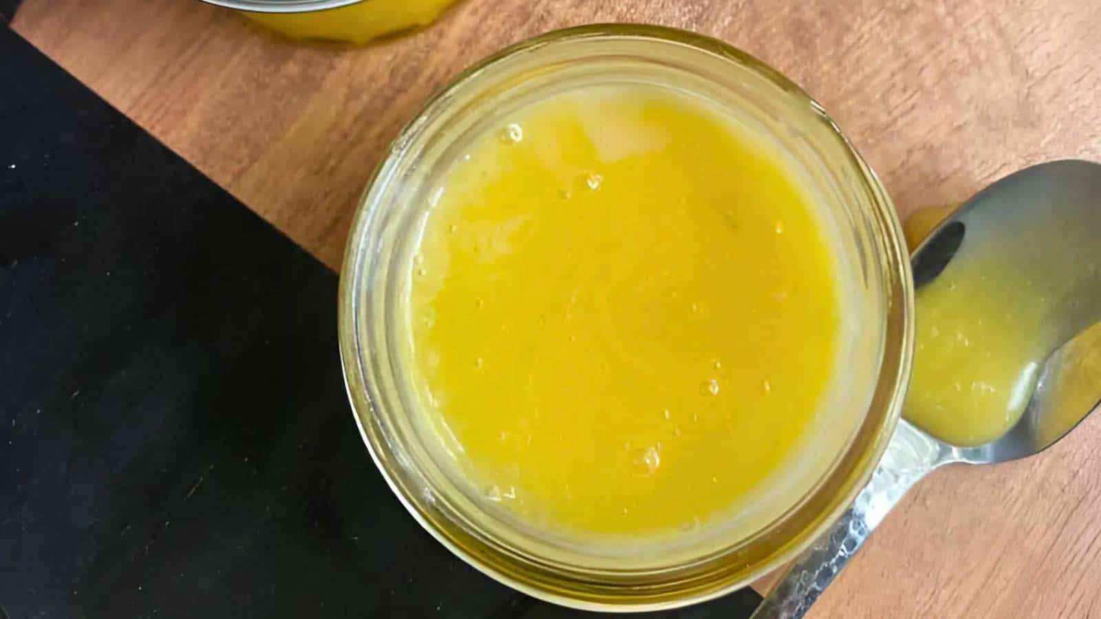 Top view of a glass jar filled with lemon curd, placed on a wooden surface next to a metal spoon that has a bit of the sauce on it.