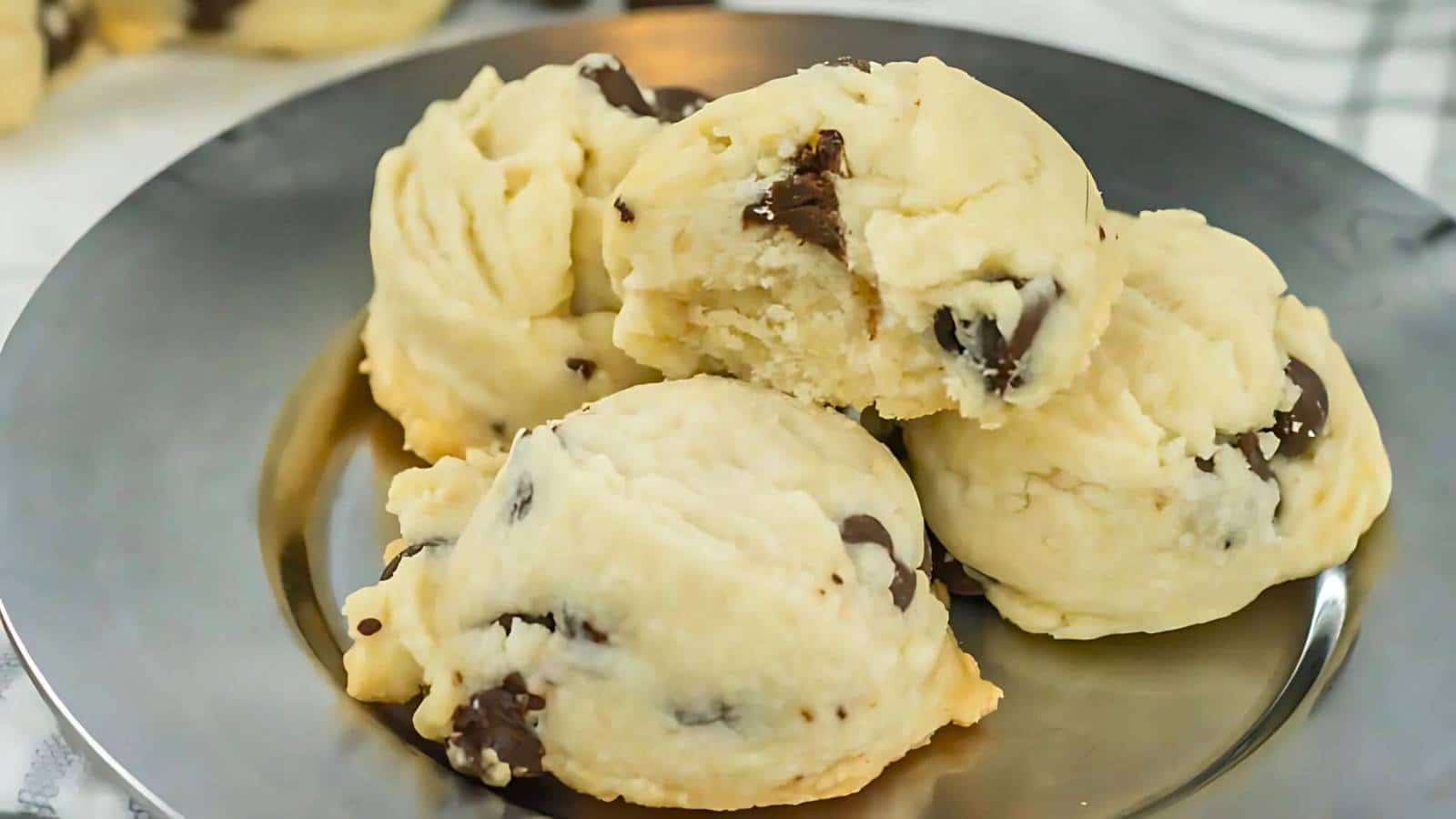 Four chocolate chip cookies are stacked on a silver plate, with one cookie showing a bite taken out.