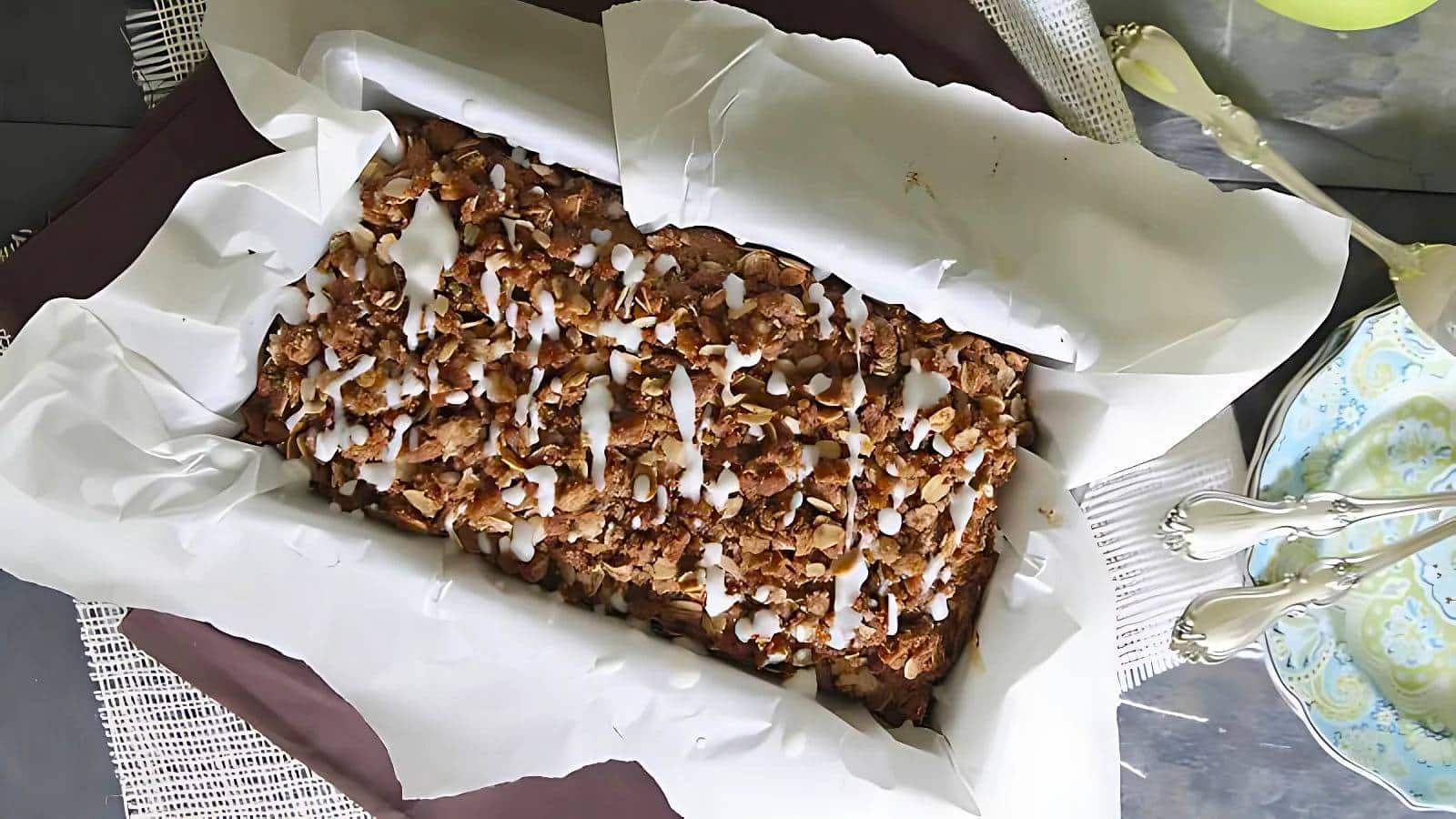 A loaf of bread topped with a drizzle of glaze sits in a parchment-lined baking dish on a table set with silverware and ornate plates.