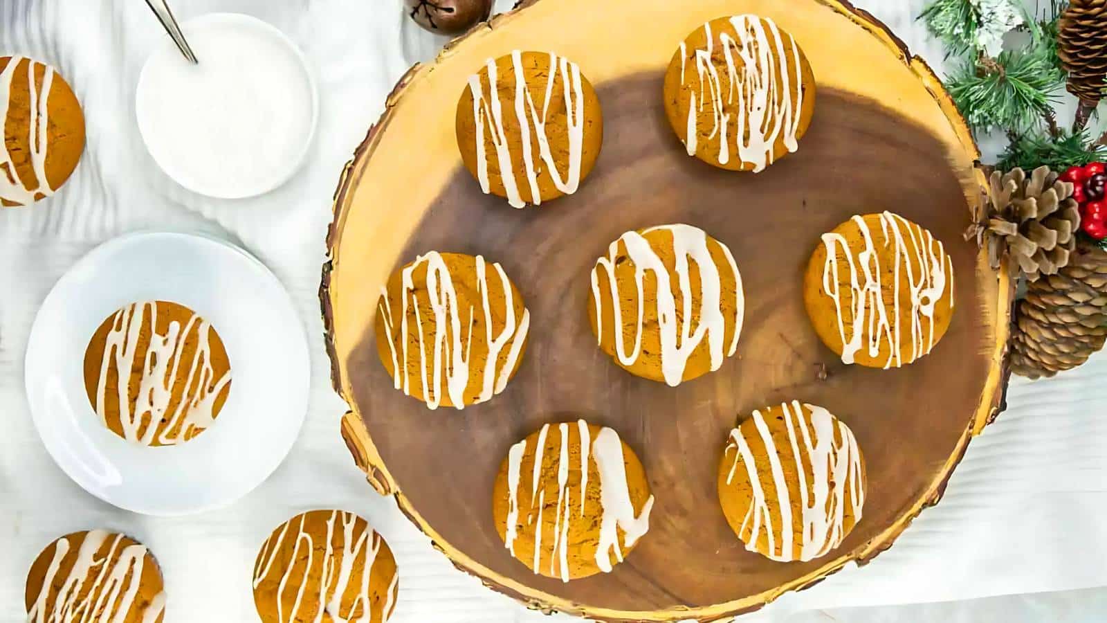 Wooden platter with nine round cookies drizzled with white icing, surrounded by pinecones and evergreen branches. A bowl and spoon are on the side.