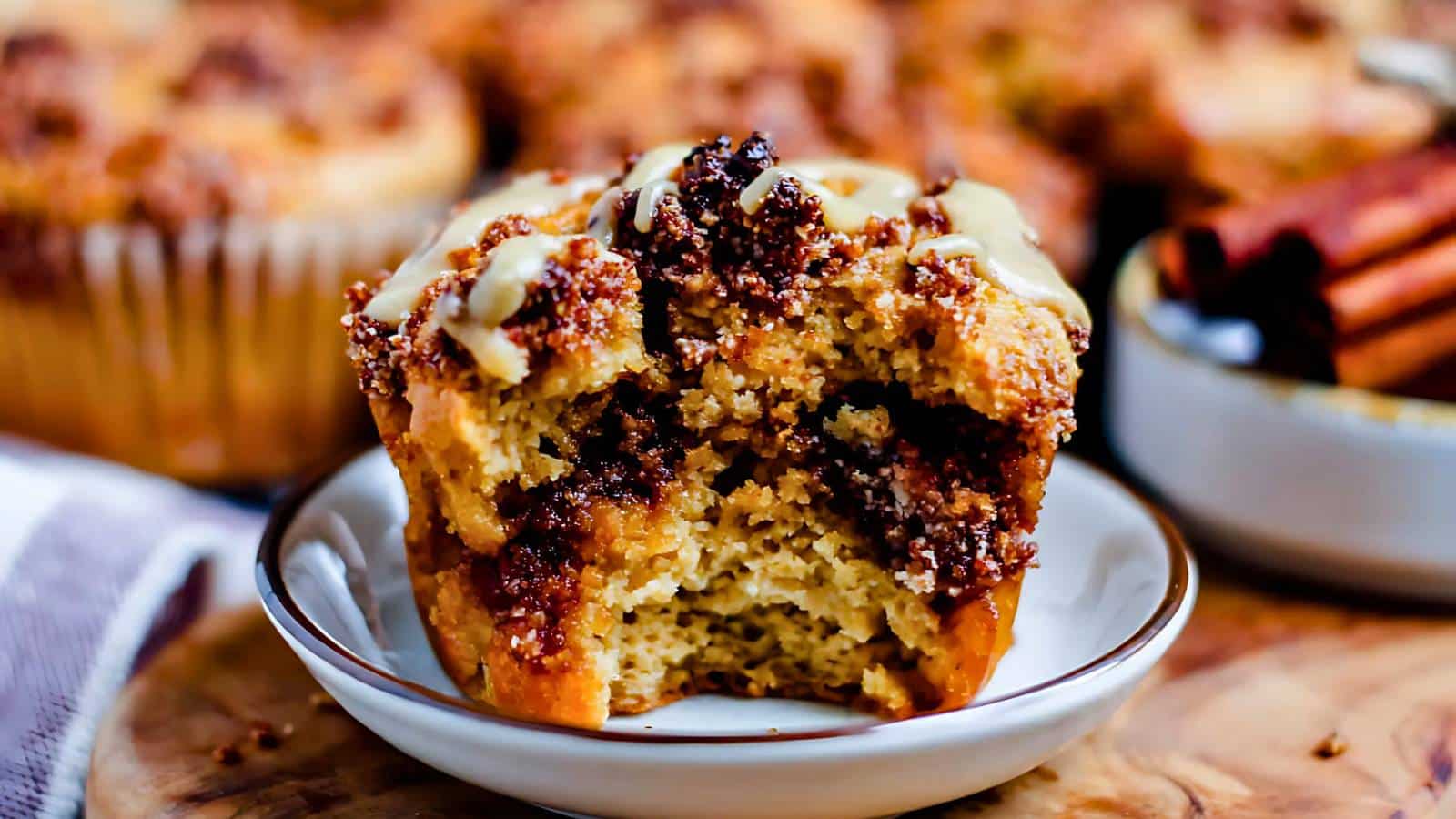 A close-up of a partially eaten muffin on a plate, topped with glaze and crumbly pieces, with cinnamon sticks in the background.