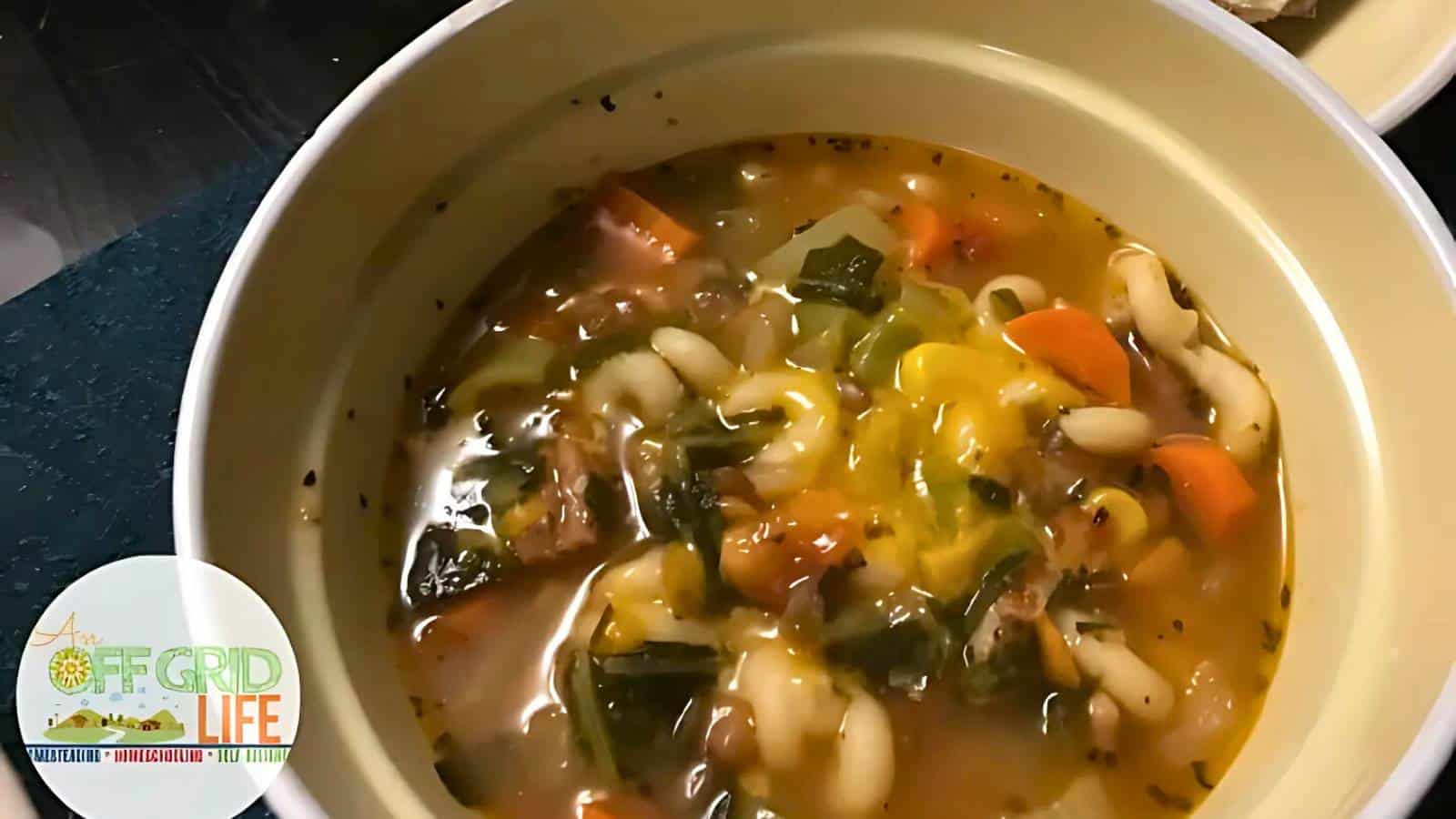 A bowl of vegetable soup with pasta, carrots, greens, and other vegetables. A logo in the corner reads "Off Grid Life.