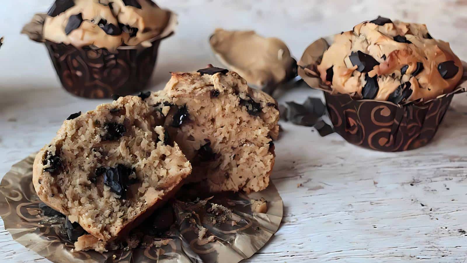 Two muffins with chocolate chips and decorative wrappers sit on a table. One muffin is whole, and the other is cut in half, revealing its interior.