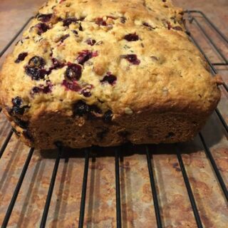 A loaf of baked berry bread with visible blueberries rests on a cooling rack atop a kitchen counter.