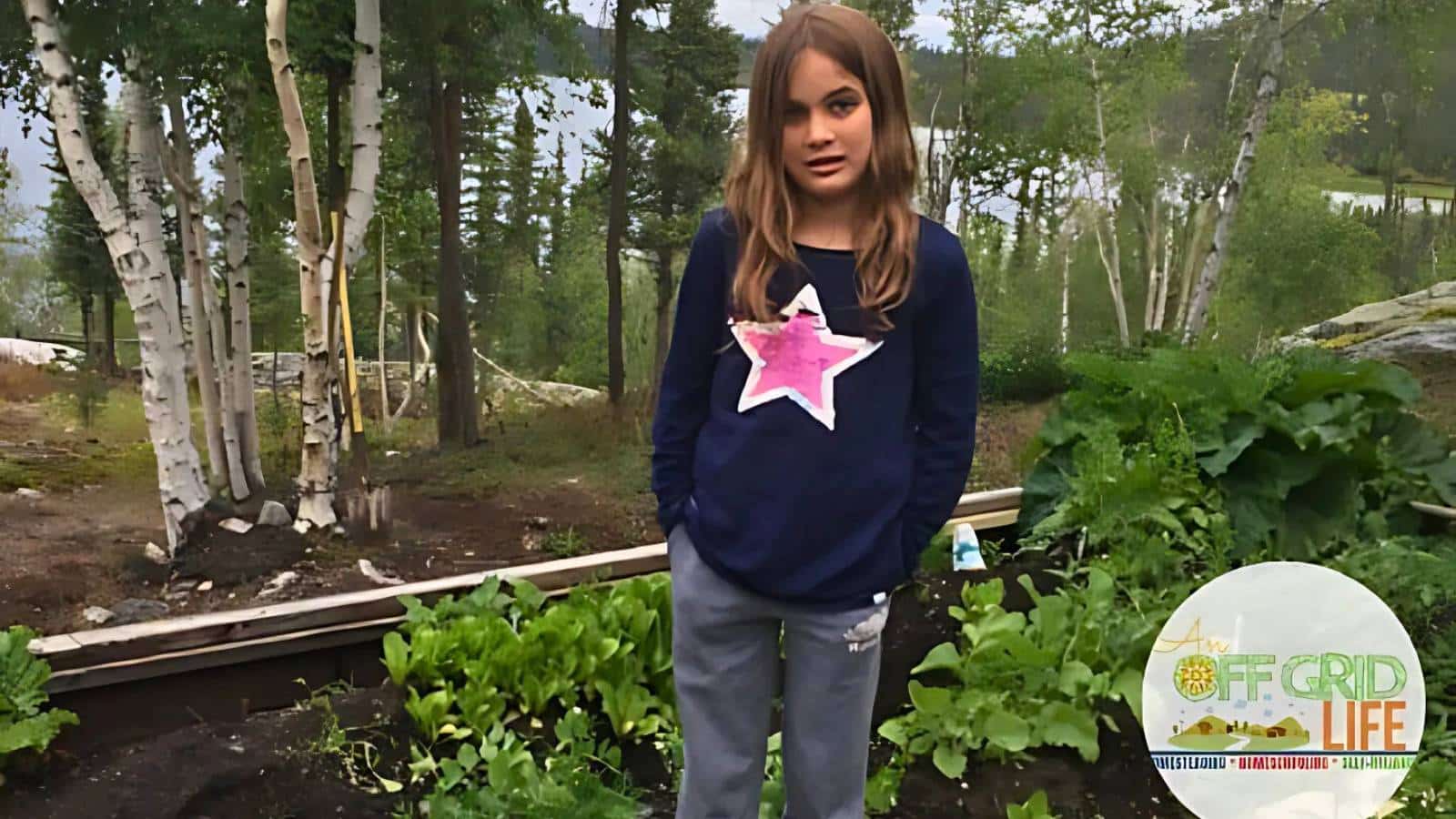 A girl stands in a garden, wearing a navy sweater with a star and gray pants. Green plants grow around her, and trees are visible in the background.