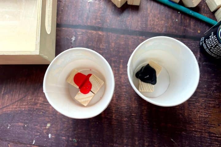 Two paper cups on a wooden surface, each containing a wooden block with red paint in one cup and black paint in the other. A partially visible wooden object is on the left.