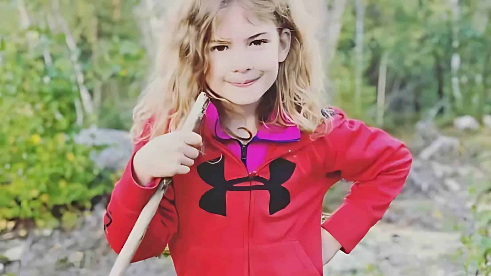 A young girl with long hair in a red jacket holds a stick while standing outdoors in a forested area.