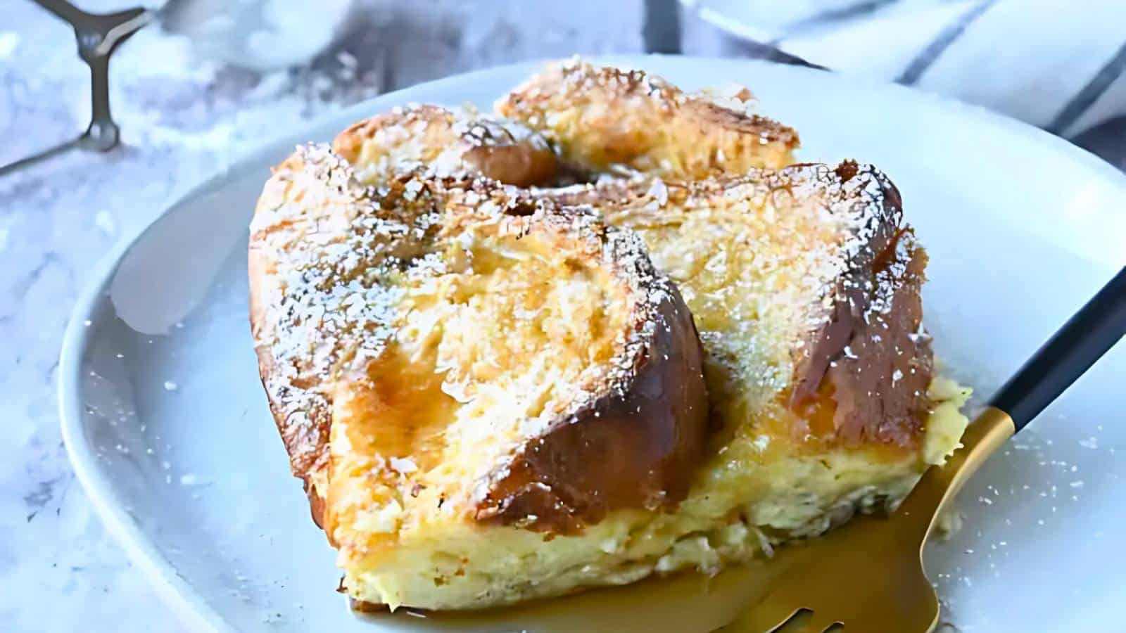A slice of French toast casserole sprinkled with powdered sugar on a white plate, with a fork resting beside it.