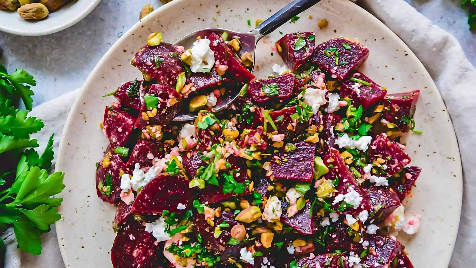 A plate of beet salad with pistachios, feta cheese, and parsley, served with a metal spoon.