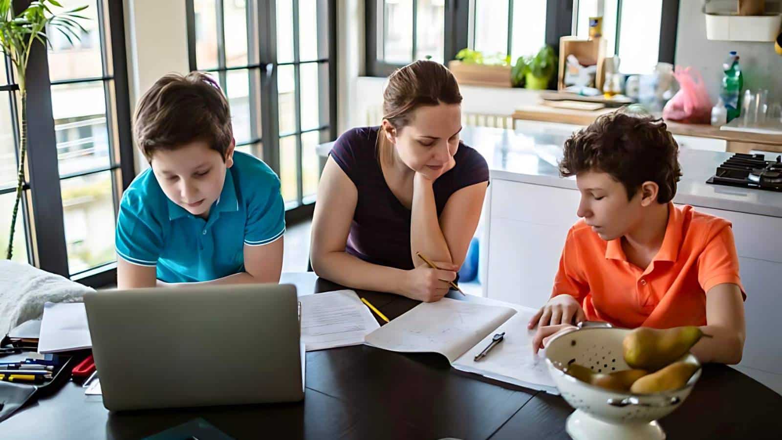 A woman helps two boys with their homework at a kitchen table. A laptop and various stationery items are on the table.