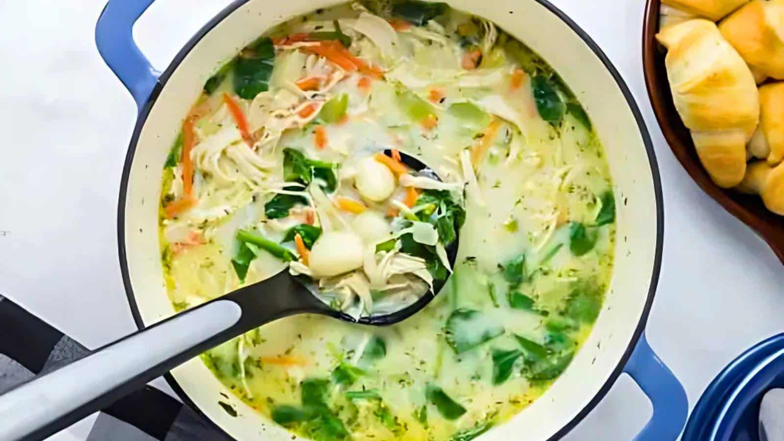 A pot of chicken soup with vegetables and noodles, featuring a ladle scooping up a portion. Crescent rolls are visible on the side.